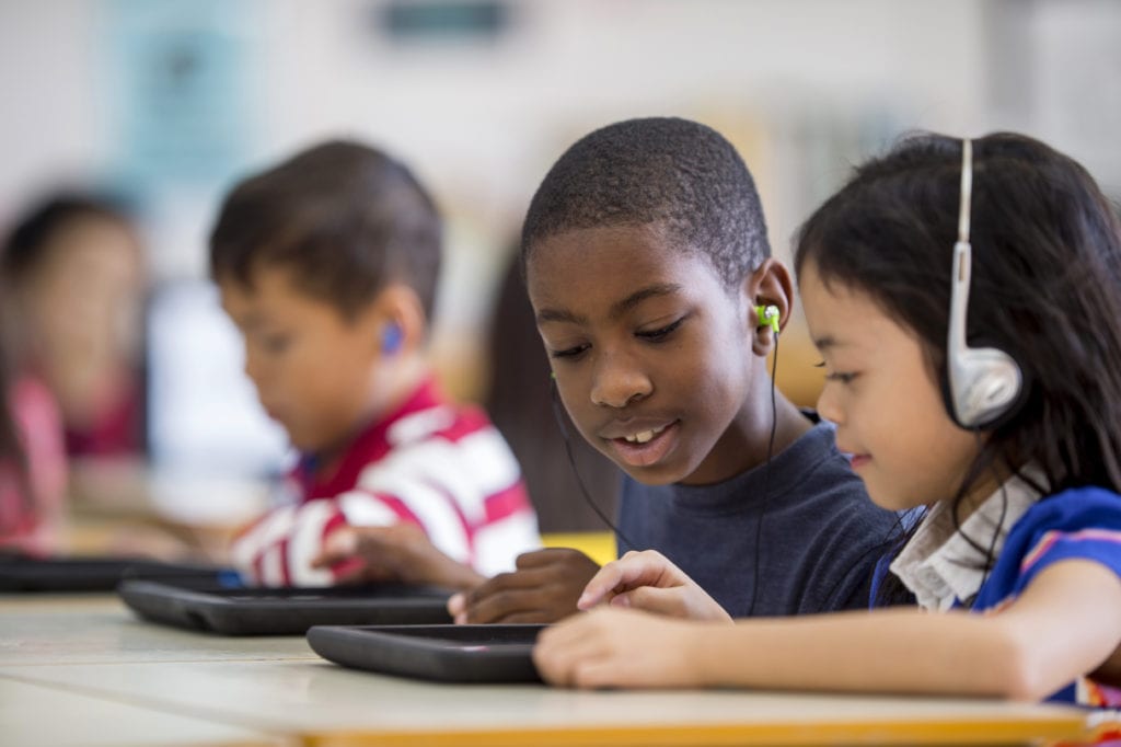 A multi-ethnic group of children are indoors in an Kids 'R' Kids classroom. They are wearing casual clothing. They are in technology class. A boy and girl are using tablet computers with headphones.
